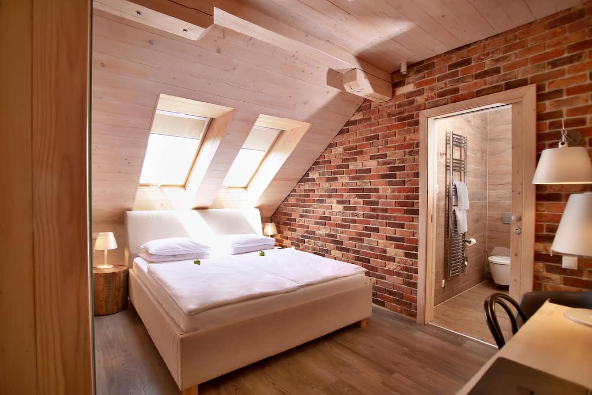 A bedroom with a sloped ceiling, a brick wall, a wooden bed, and a small table with a chair.