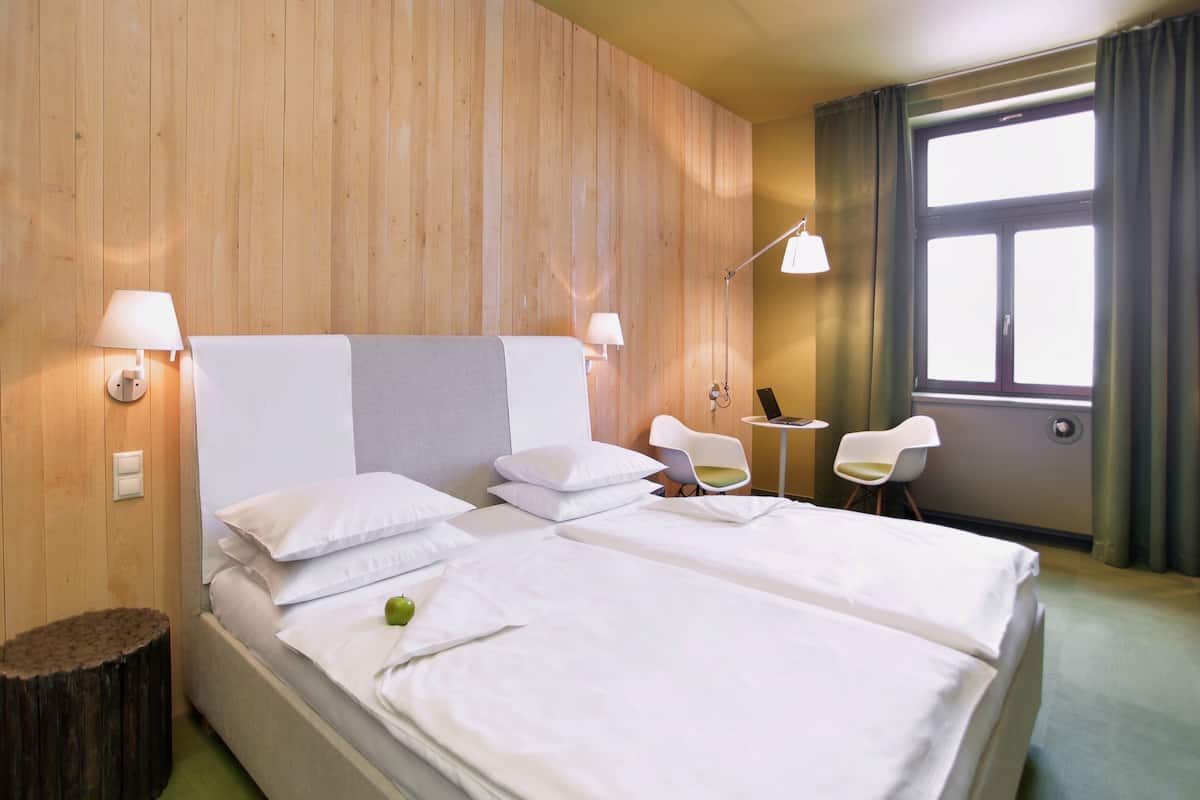 A hotel room with a wooden wall, a large bed with white bedding, two bedside tables with lamps, a small round table with a chair, and a window with curtains.