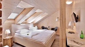 Un dormitorio moderno con una cama grande, un escritorio y una silla. Hay tragaluces en el techo, un televisor de pantalla plana montado en la pared y una pequeña mesa con una lámpara.