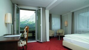 A hotel room with a bed, a desk, a chair, and a view of mountains through a glass door. - Sorell Hotel Asora (Arosa)