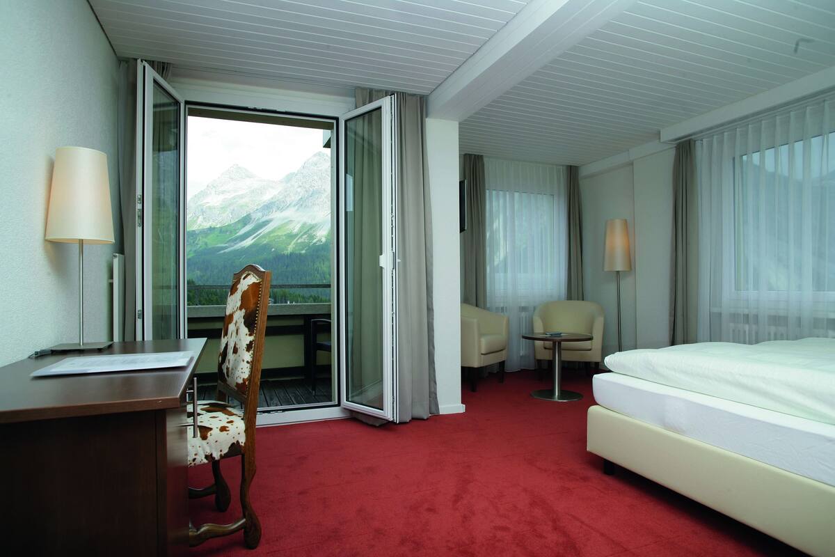 A hotel room with a bed, a desk, a chair, and a view of mountains through a glass door.