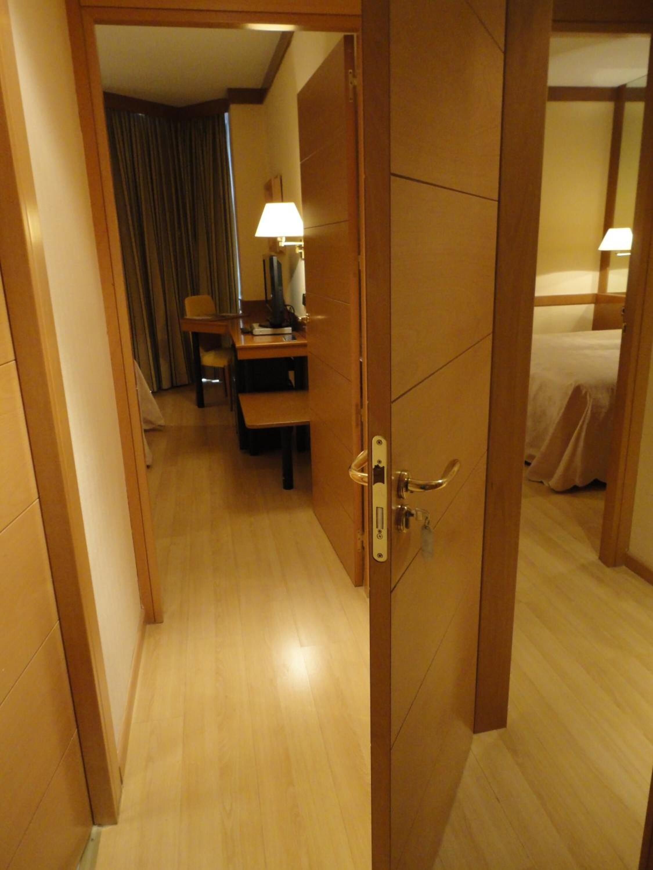 A hotel room with a wooden door, a desk with a lamp, a bed, and a curtain.