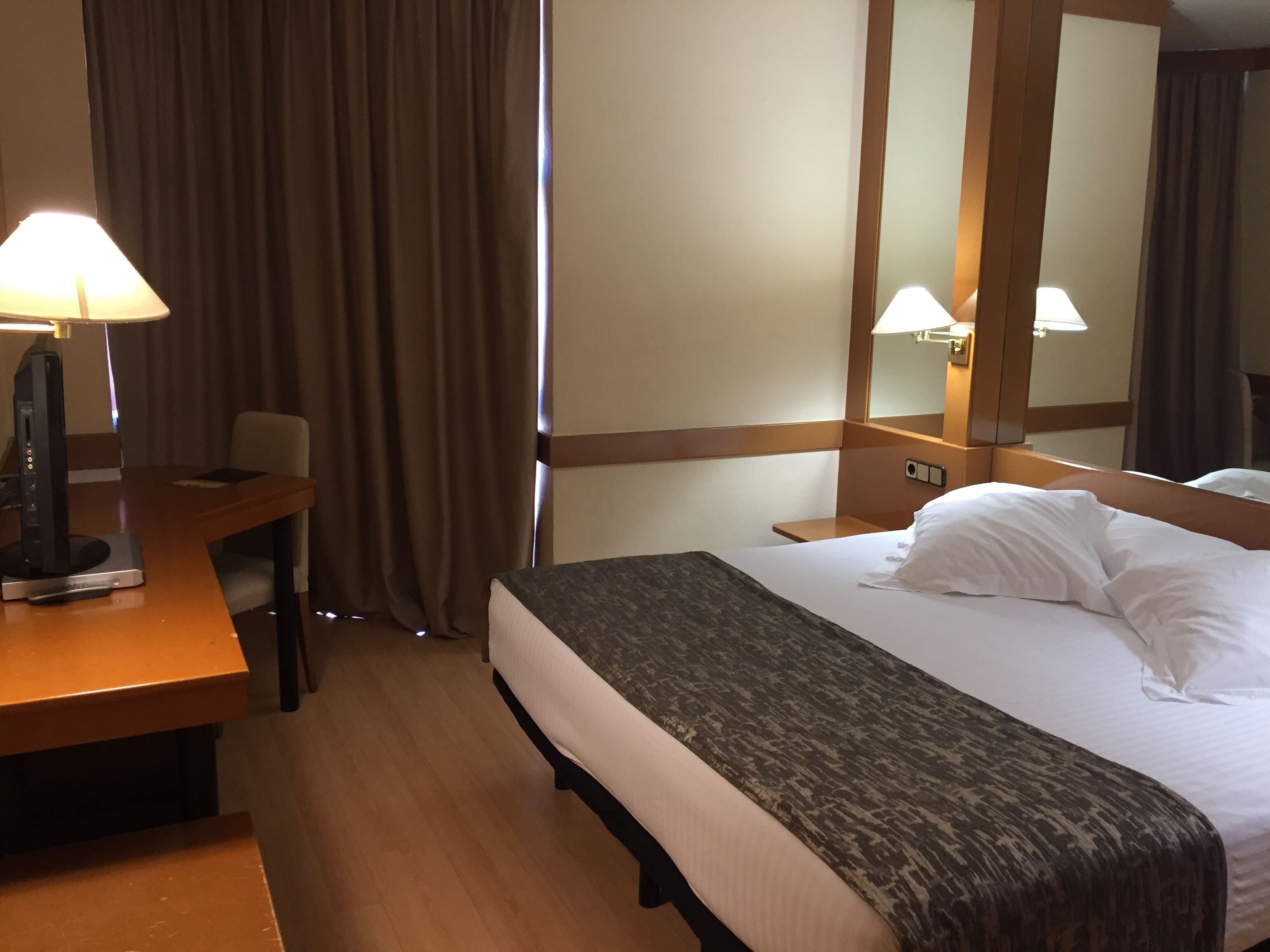 Ett hotellrum med en säng, ett skrivbord, en stol, en lampa och en spegel.