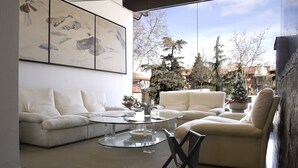A modern living room with two white sofas, a glass coffee table, and a large window offering a view of trees and buildings. - Hotel AR Los Arcos (Segovia)