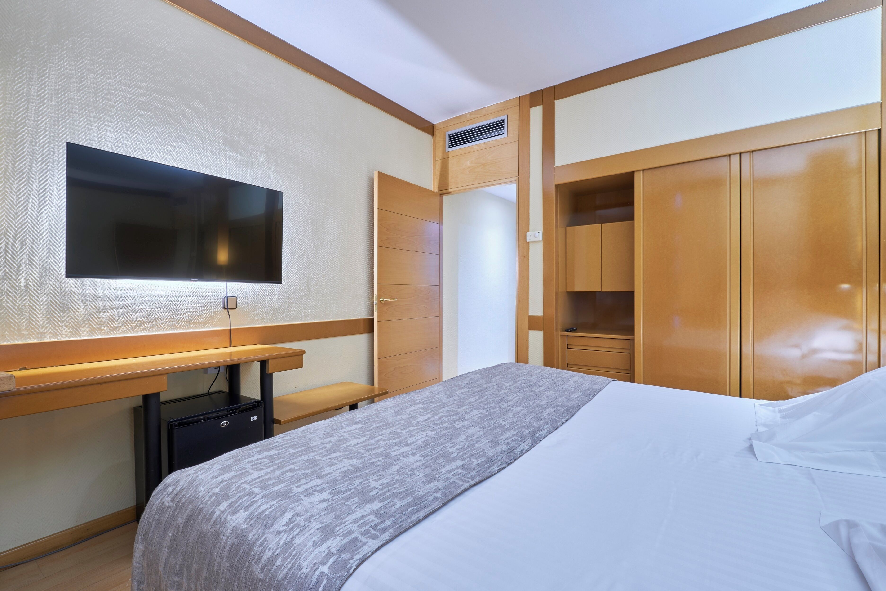 Habitación de hotel con cama, escritorio con televisor de pantalla plana, armario y pequeño ventilador de mesa.
