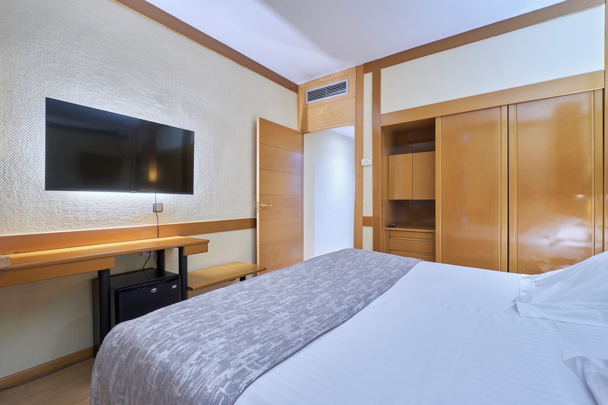 Habitación de hotel con cama, escritorio con televisor de pantalla plana, armario y pequeño ventilador de mesa.