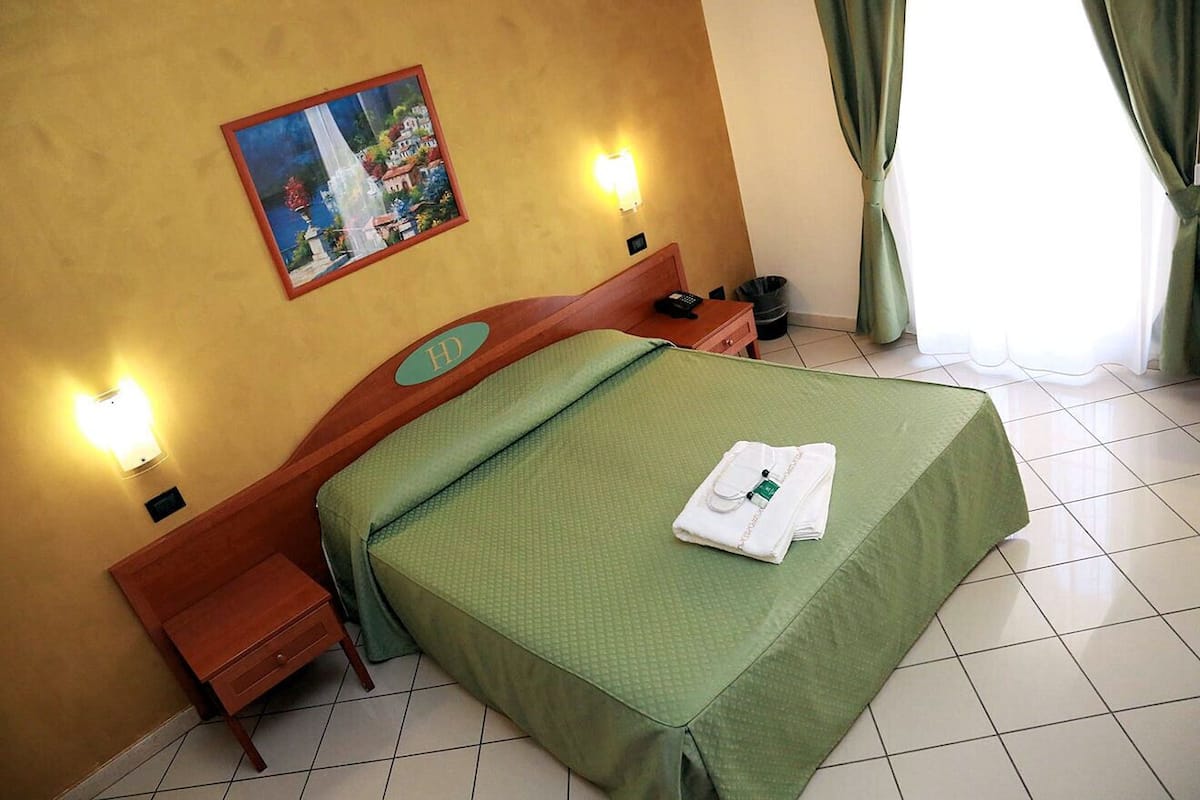 A hotel room with a bed, bedside tables, a nightstand, a lamp, a painting on the wall, and a window with curtains.
