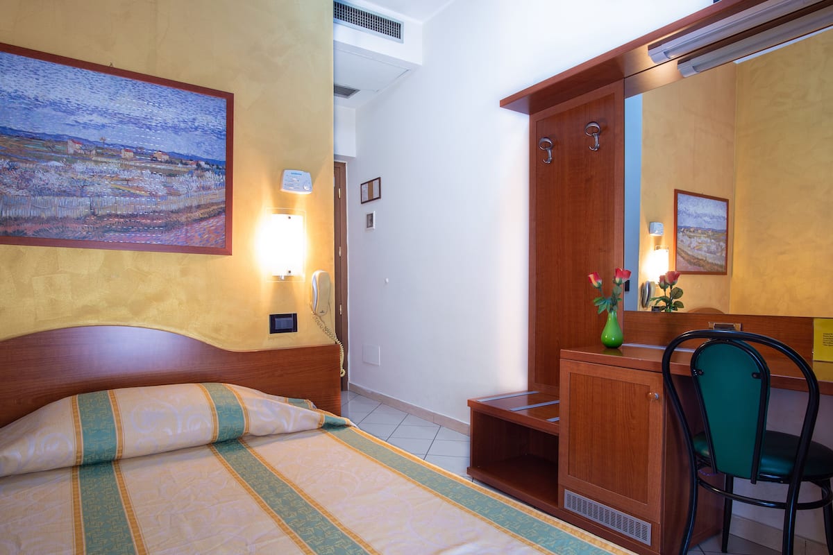 A hotel room with a bed, a desk, a chair, a mirror, and a painting on the wall.