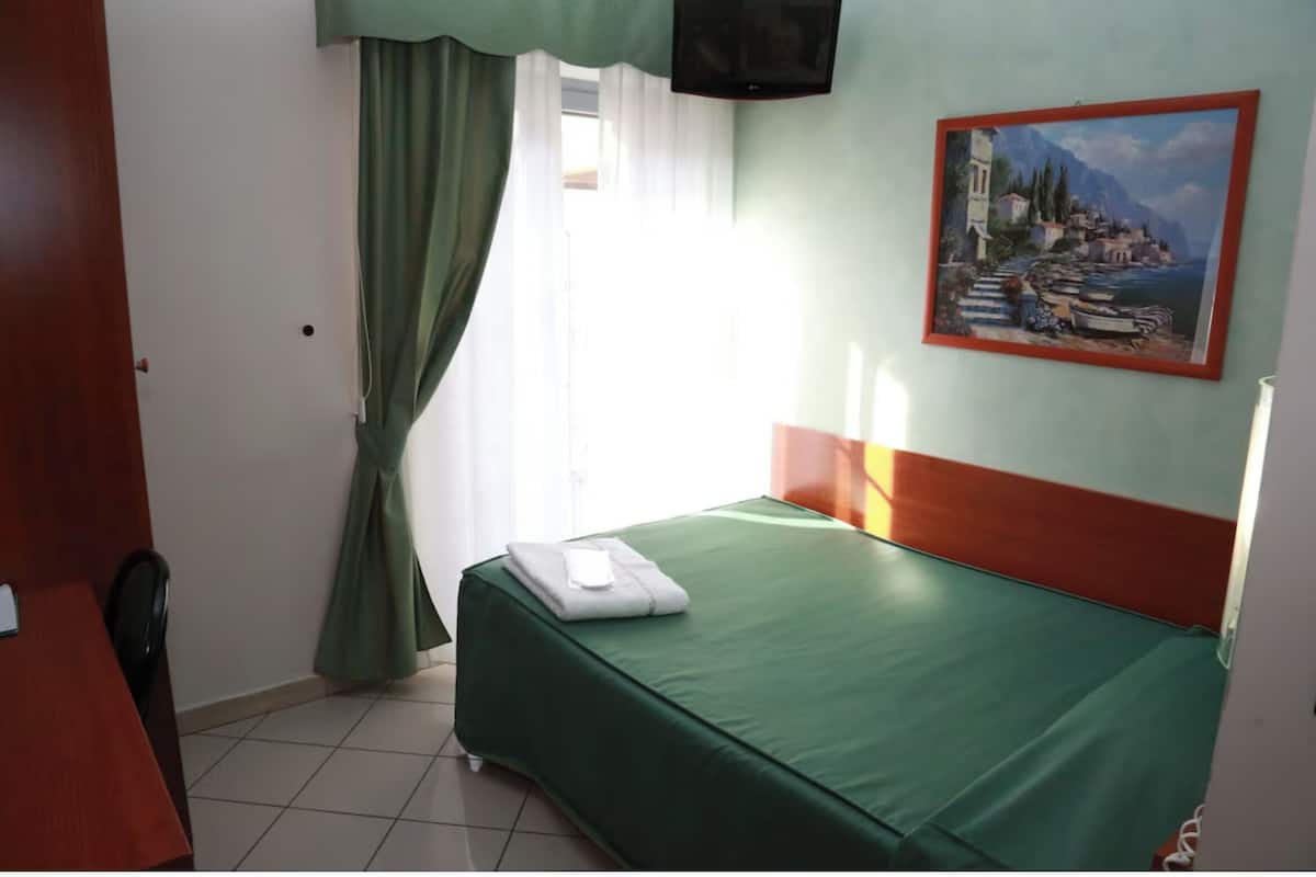 A room with a bed, a desk, a chair, a painting, and a window with curtains.