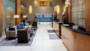 Een hotel lobby met een receptiebalie, een zithoek met sofa’s en fauteuils, een trap naar een hoger gelegen niveau en decoratieve planten.
