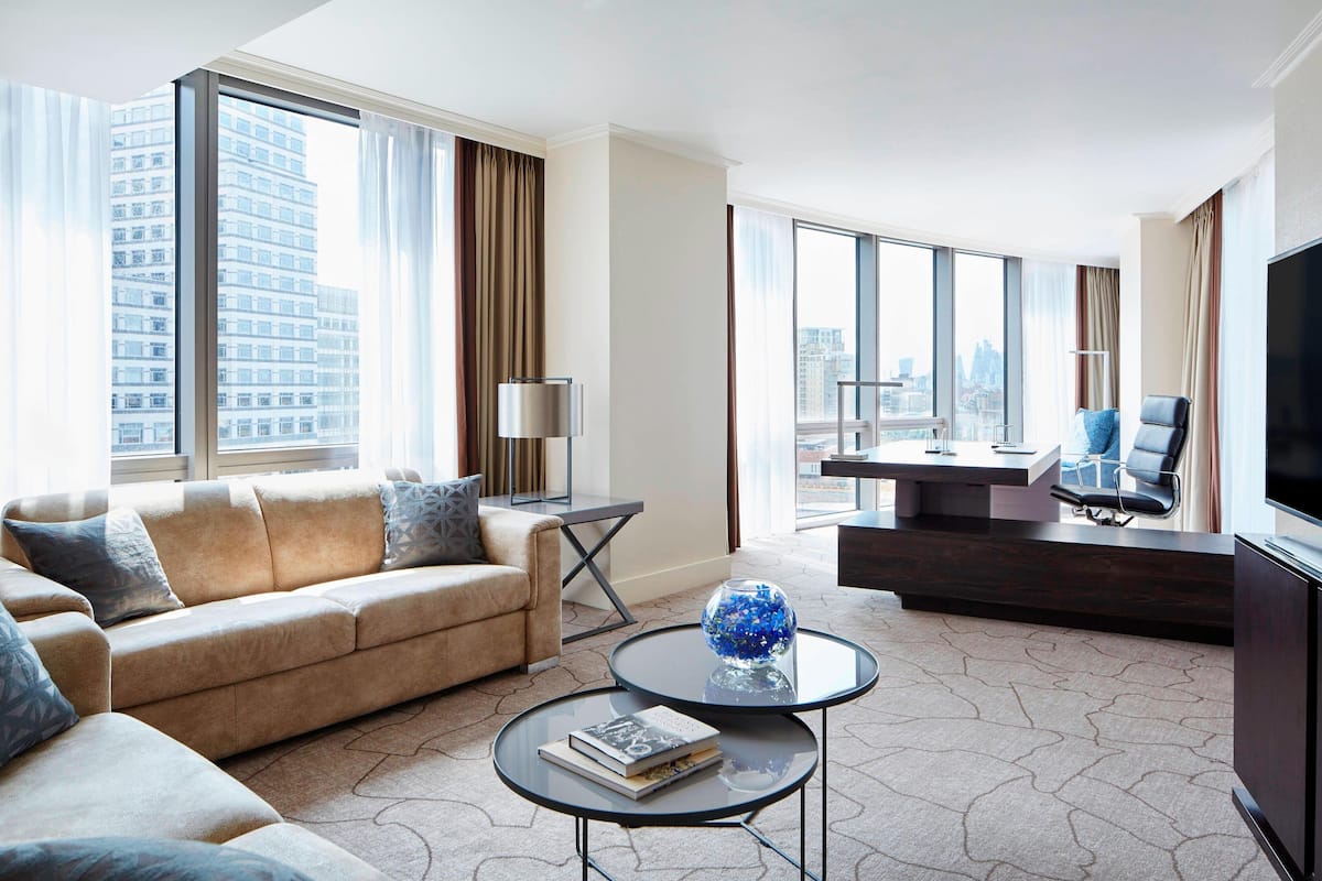 Ein modernes Hotelzimmer mit einer großen Couch, einem Couchtisch, einem Fernseher und Blick auf die Stadt durch große Fenster.