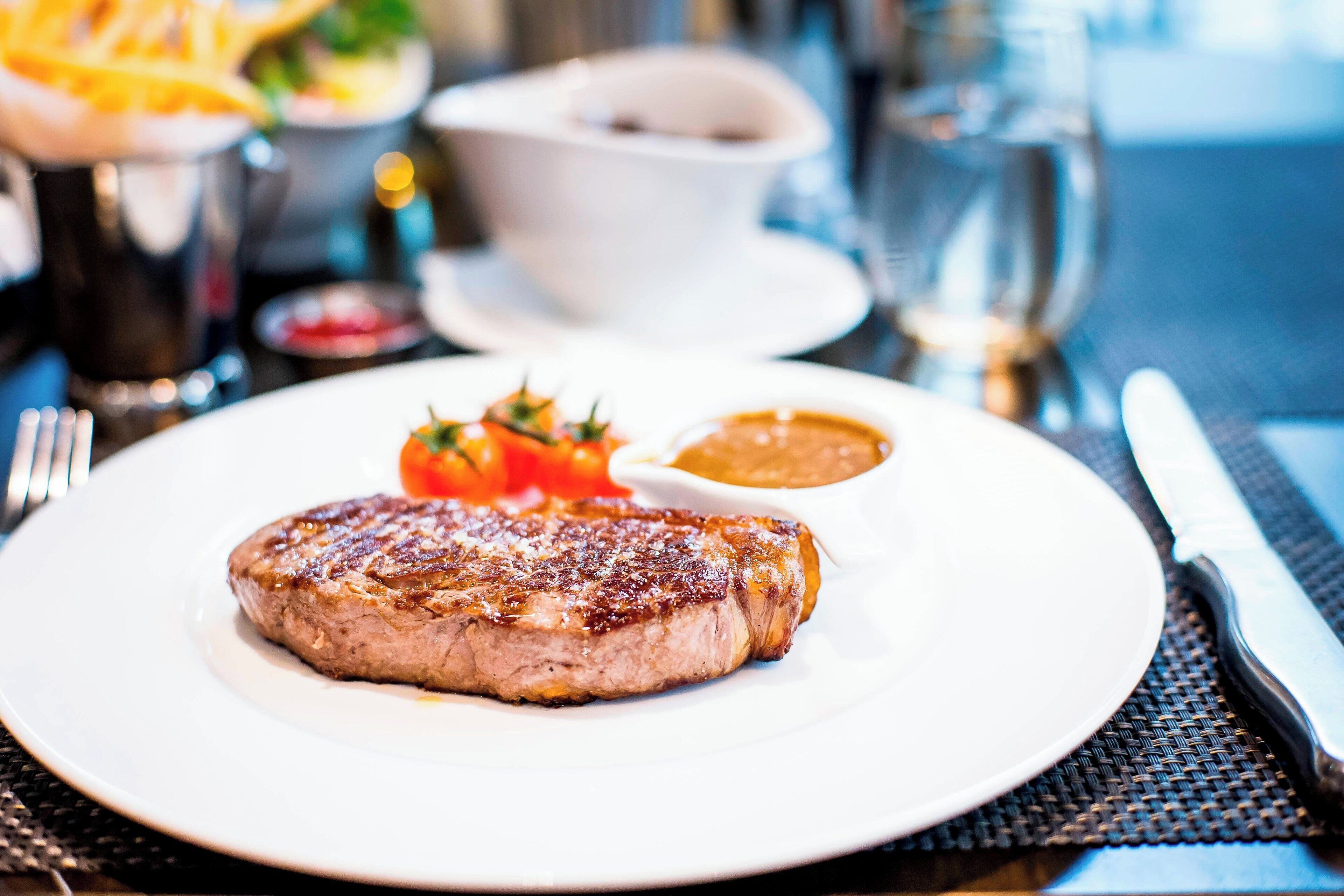 A plate with a grilled steak, a small bowl of sauce, and a garnish of tomatoes and herbs.