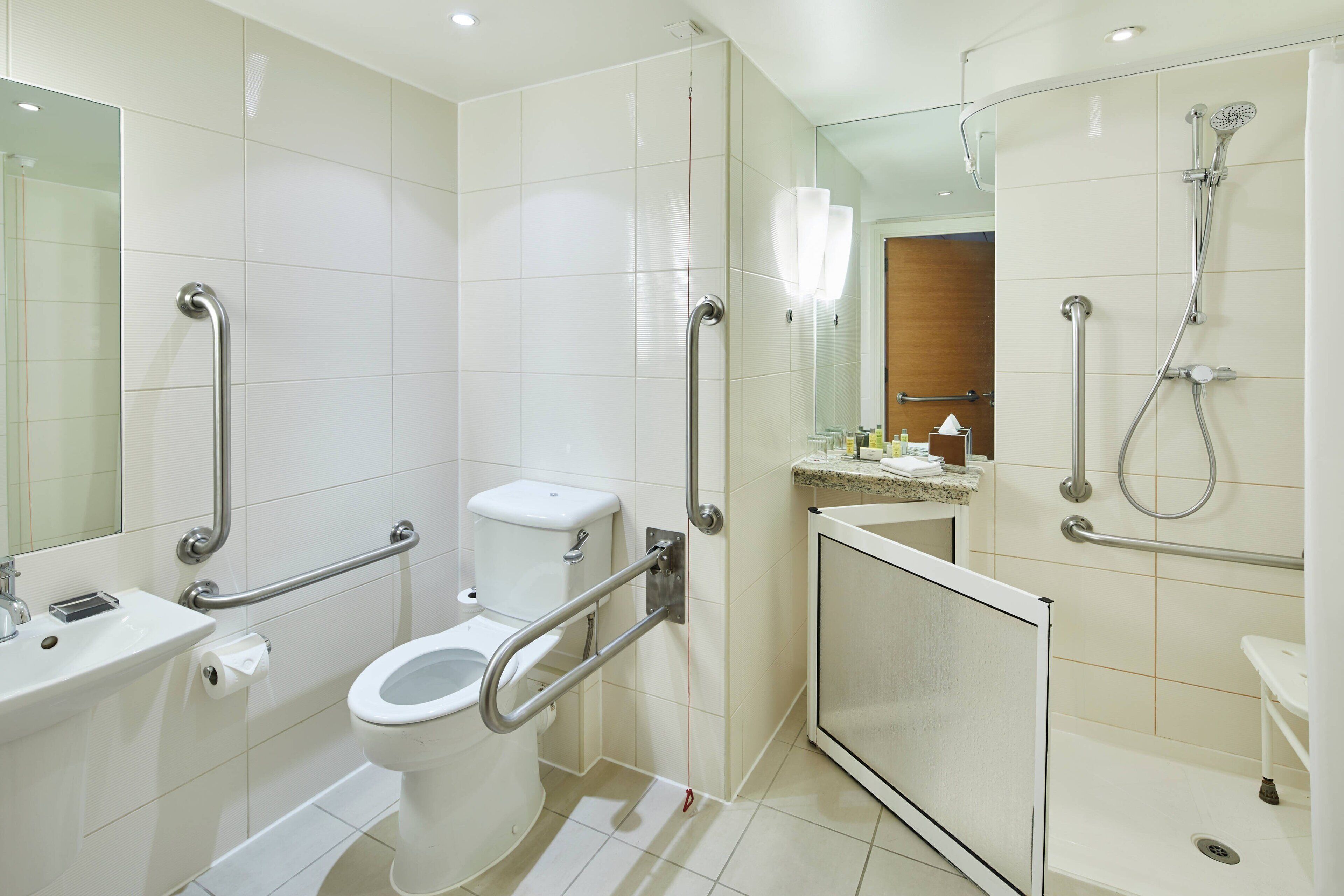 A modern bathroom with a toilet, sink, and shower. It includes grab bars and a handrail for accessibility.