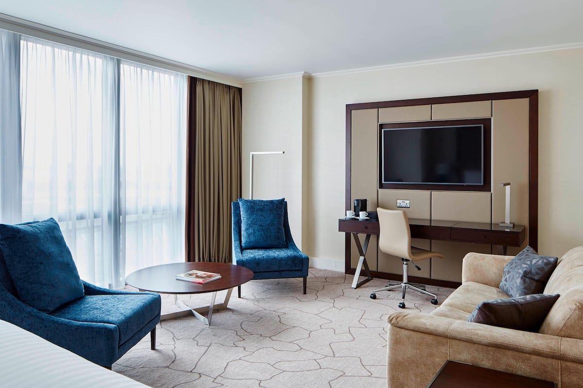 Ein Hotelzimmer mit großem Fernseher, zwei Sesseln, einer Couch, einem Couchtisch und einem Schreibtisch mit Stuhl.