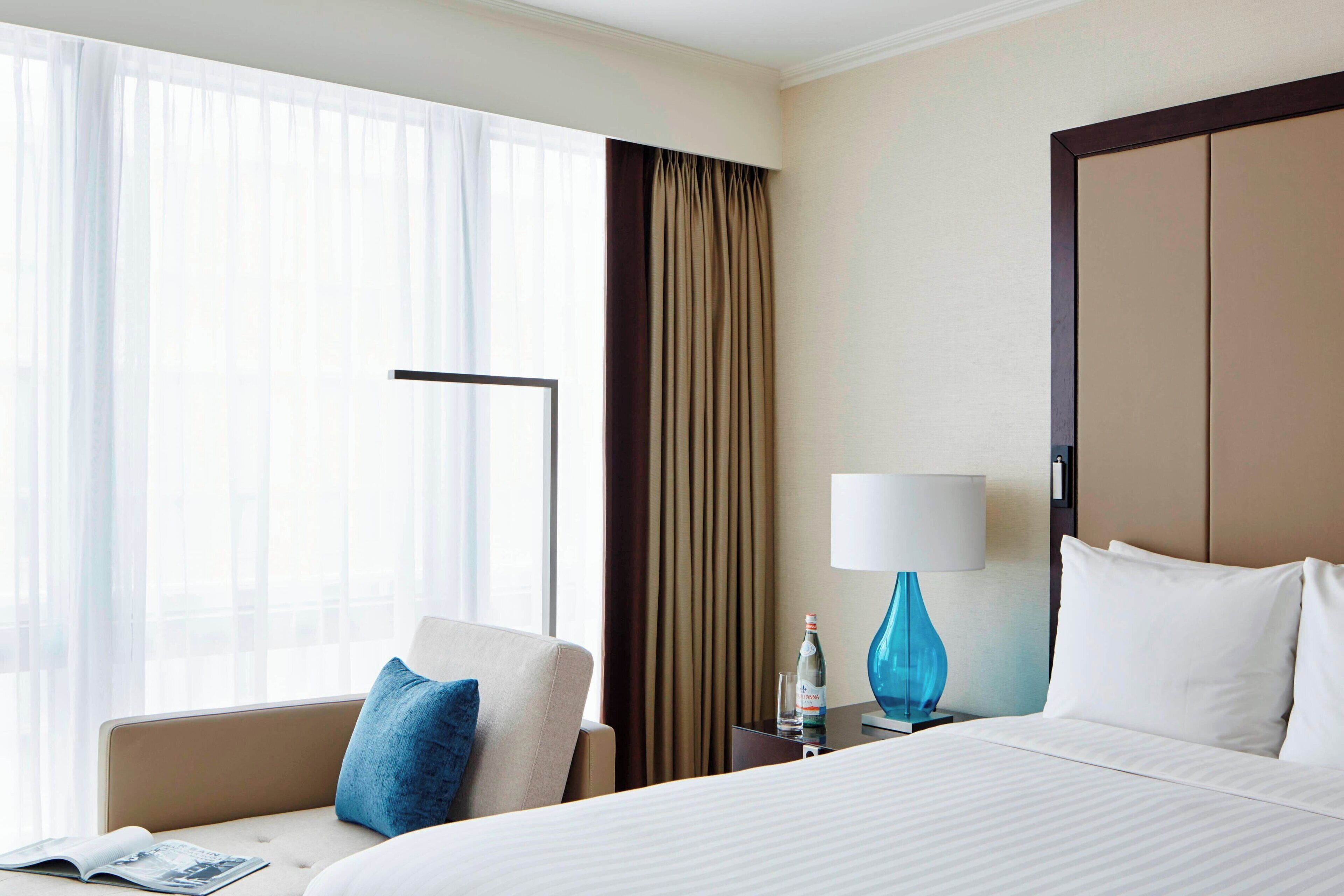 Een hotelkamer met een bed, een stoel, een nachtkastje met een lamp en een raam met gordijnen.
