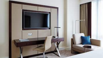 A hotel room with a large flat-screen TV mounted on a wall, a desk with a chair, a sofa with a blue pillow, and a bedside table.