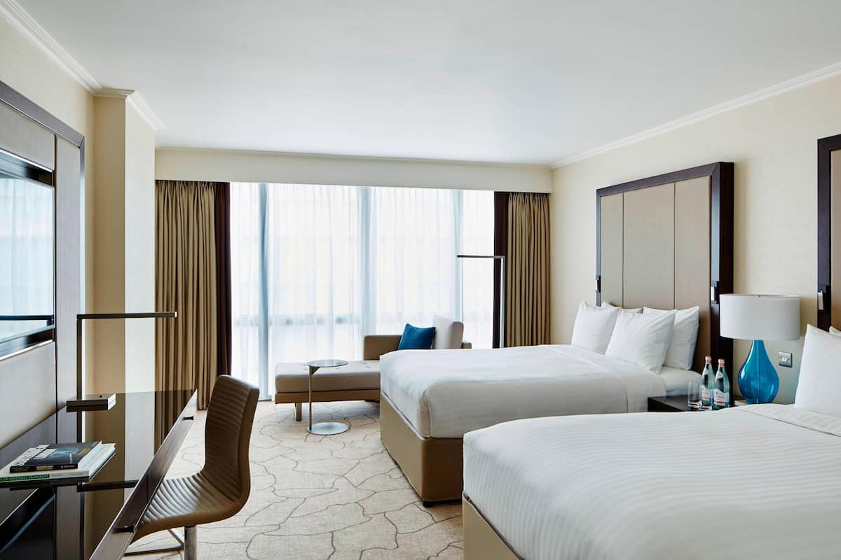 Ein Hotelzimmer mit zwei Betten, einem Schreibtisch, einem Stuhl, einem kleinen Tisch und einem großen Fenster mit Vorhängen.