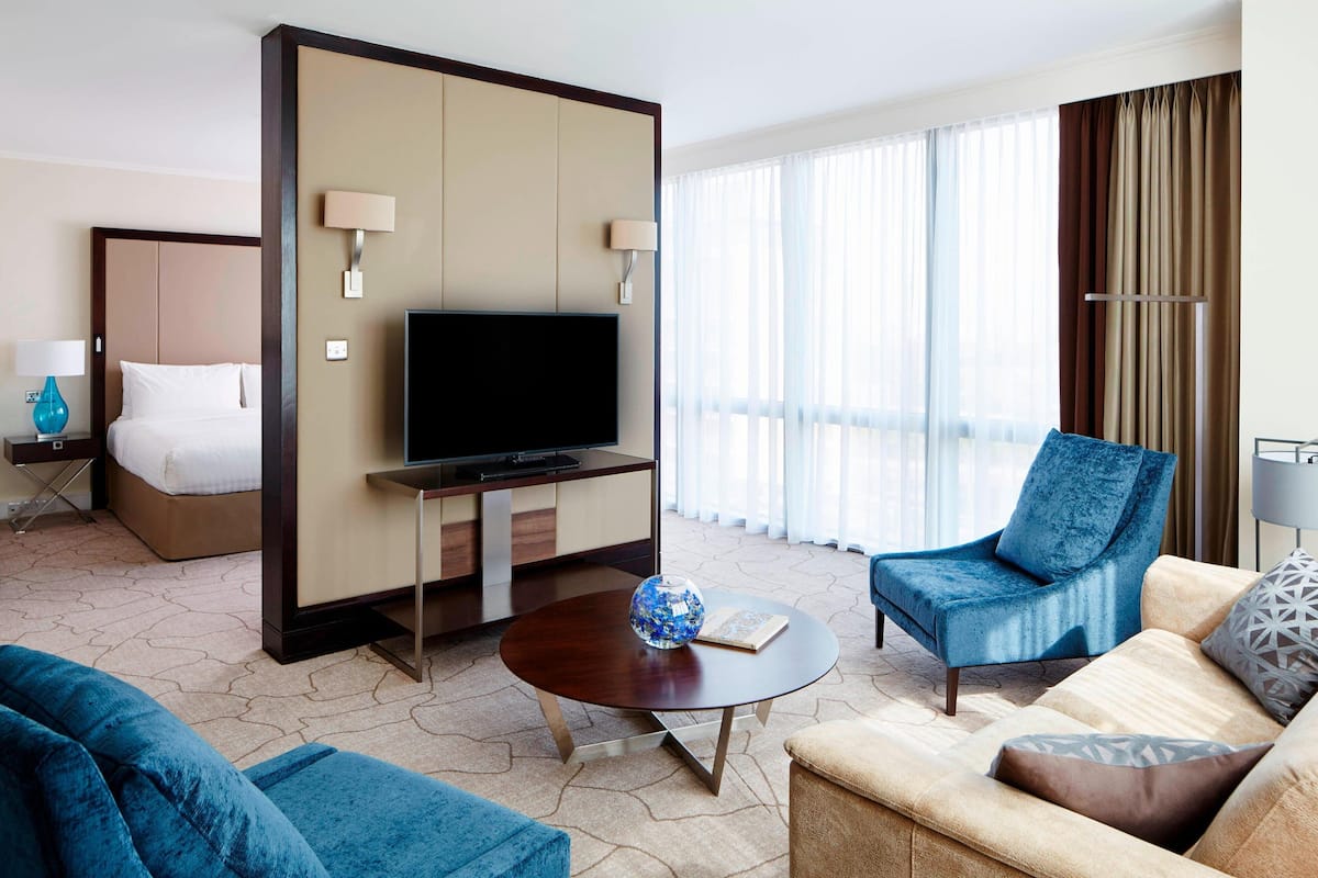 Ein Hotelzimmer mit einem Bett, einem Fernseher, einem Couchtisch mit einem dekorativen Gegenstand, einem blauen Sessel und einem Sofa.