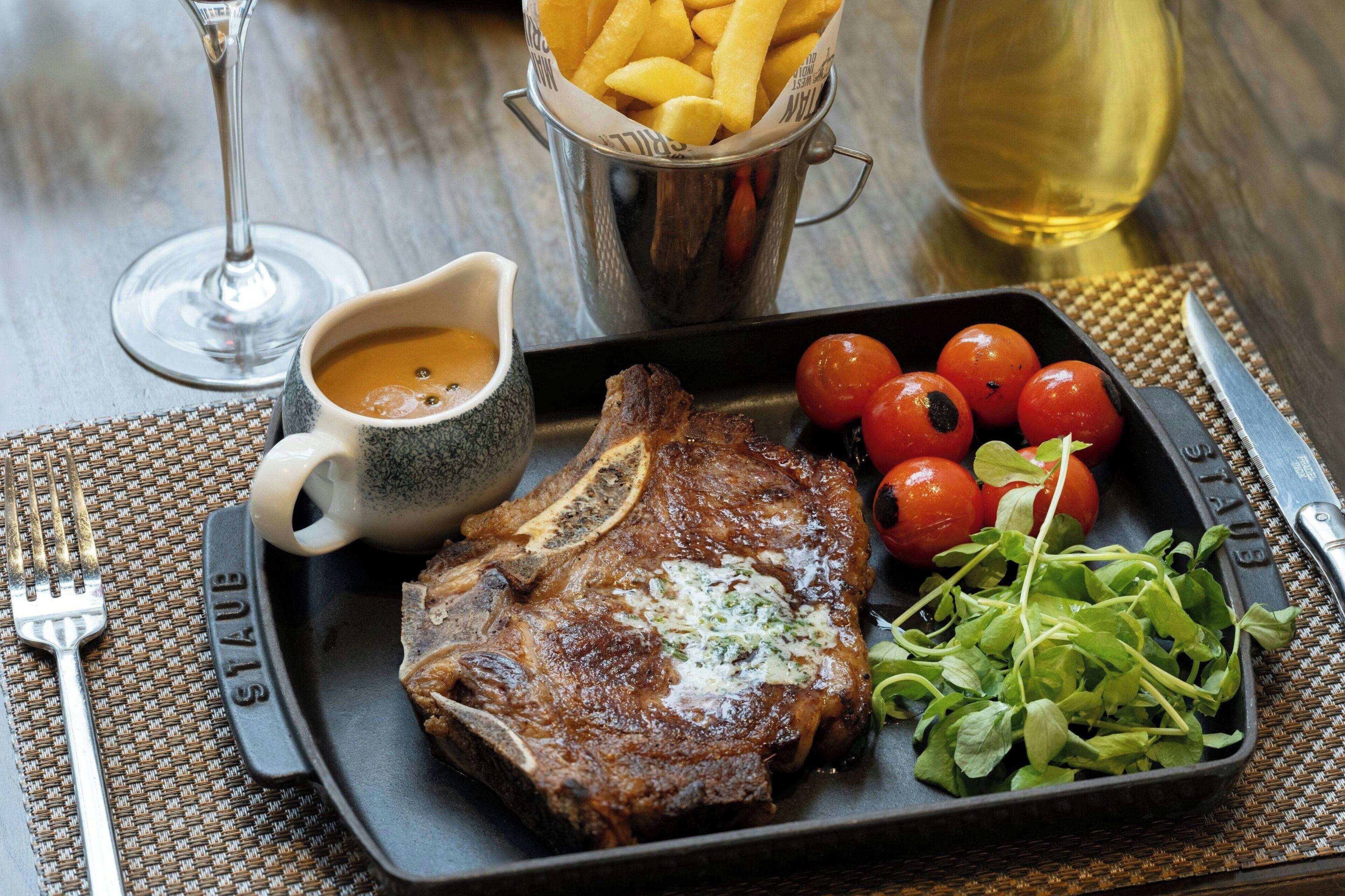 Un plato de carne con tomates cherry y verduras, una copa de vino y una cesta de papas fritas sobre una mesa.