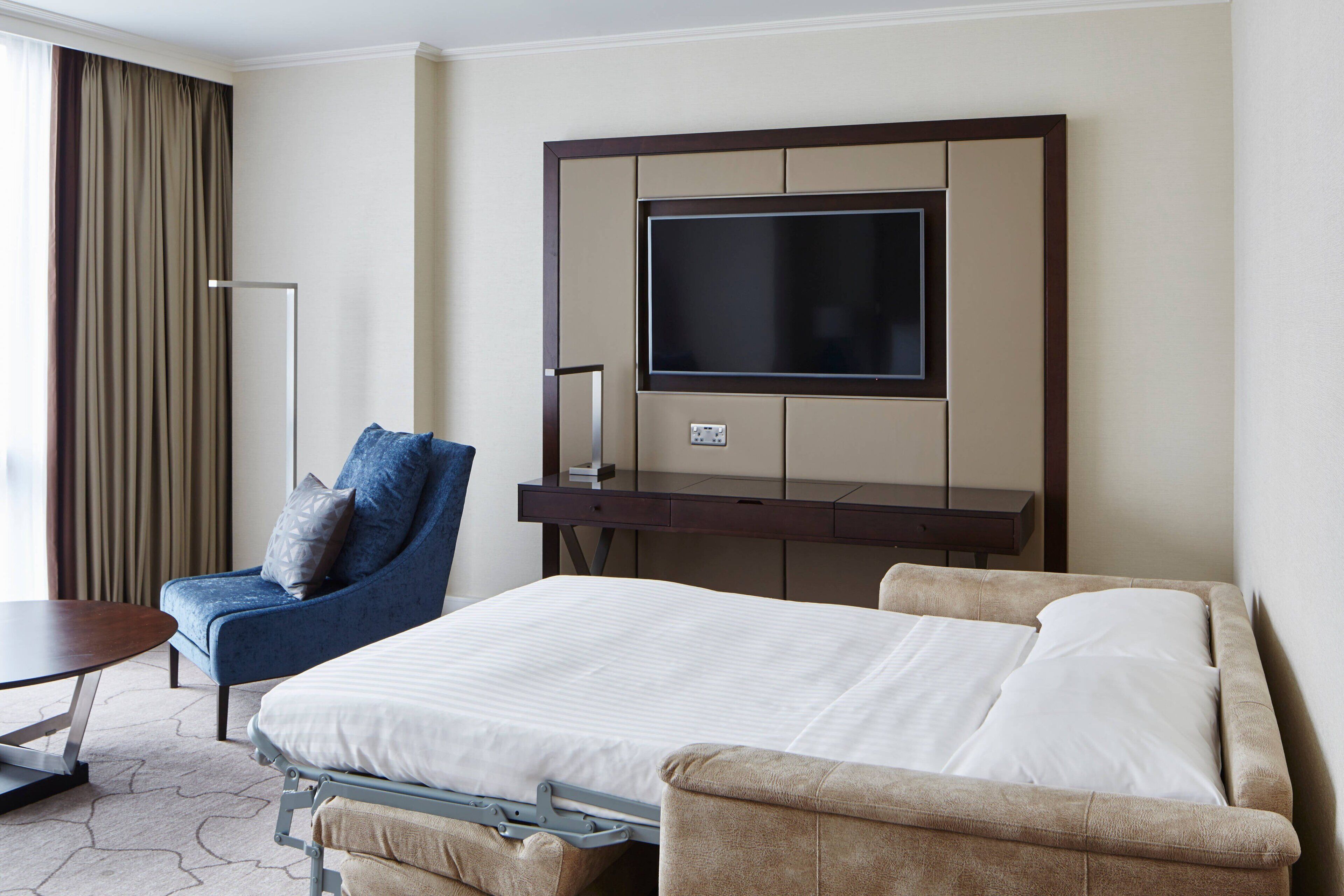 Een hotelkamer met een bed, een stoel, een bureau en een aan de muur bevestigde televisie.