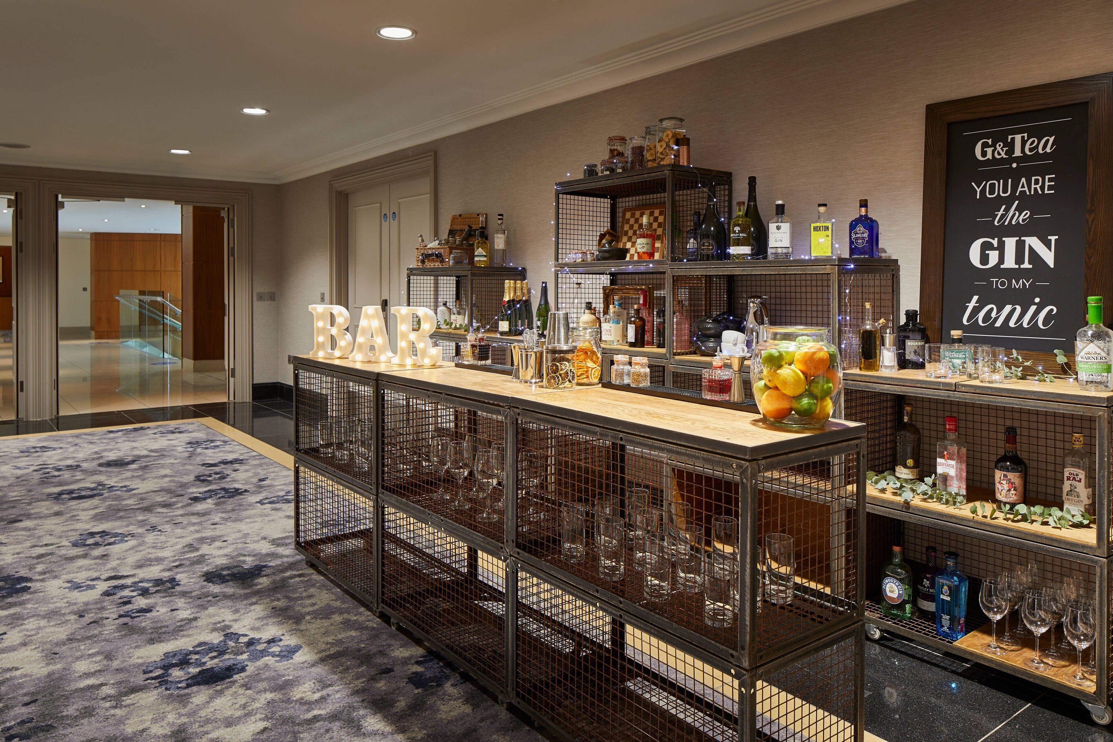 Eine Bar mit Theke, Regalen voller Flaschen, Gläsern und einem Schild mit der Aufschrift 'G&Tea you are the gin to my tonic'.