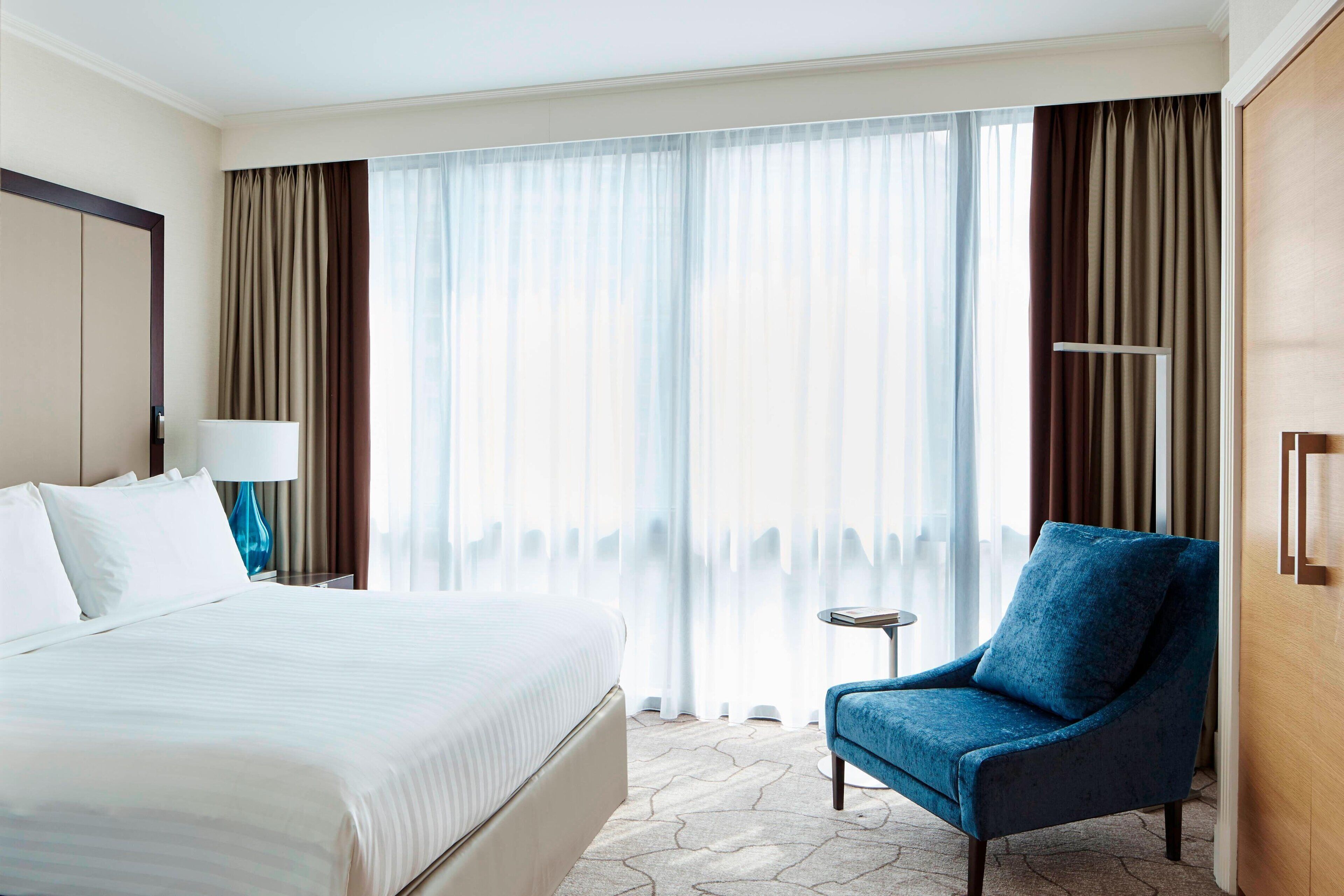 Une chambre d’hôtel avec un grand lit, un fauteuil bleu, une petite table et une grande fenêtre avec des rideaux.