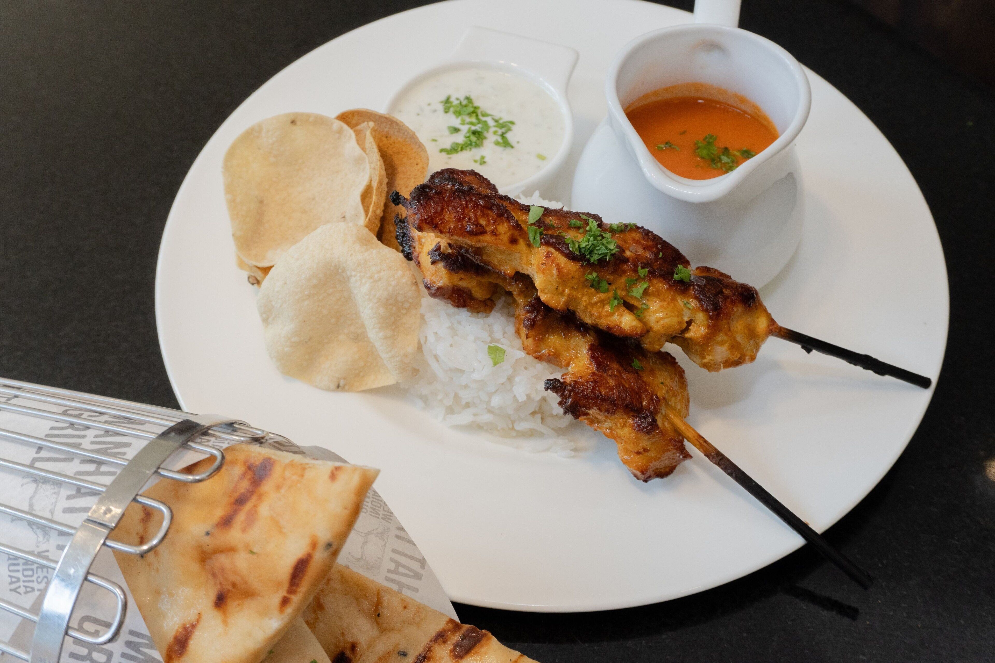 A plate with grilled chicken skewers, rice, flatbread, and two dipping sauces.
