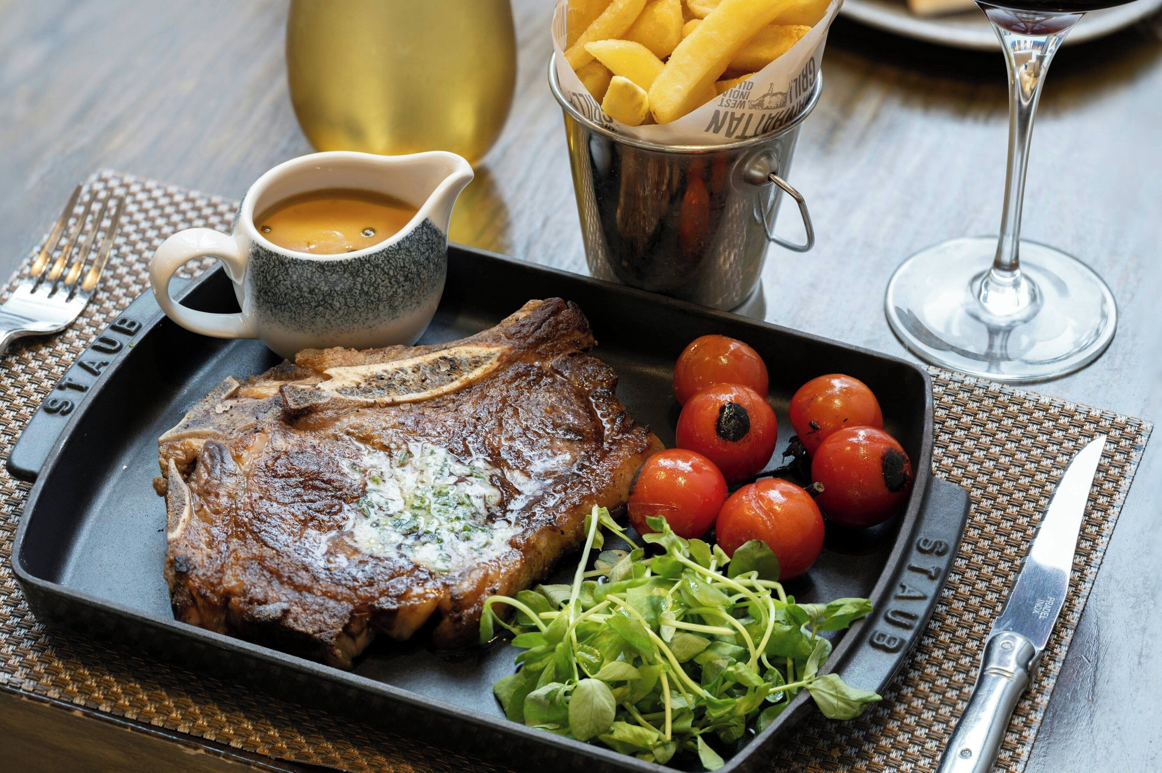 A steak with a side of cherry tomatoes and greens, a cup of sauce, and a container of fries on a table.