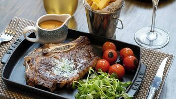 A steak with a side of cherry tomatoes and greens, a cup of sauce, and a container of fries on a table.