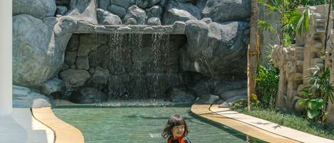Niño en una piscina con cascada y rocas al fondo.