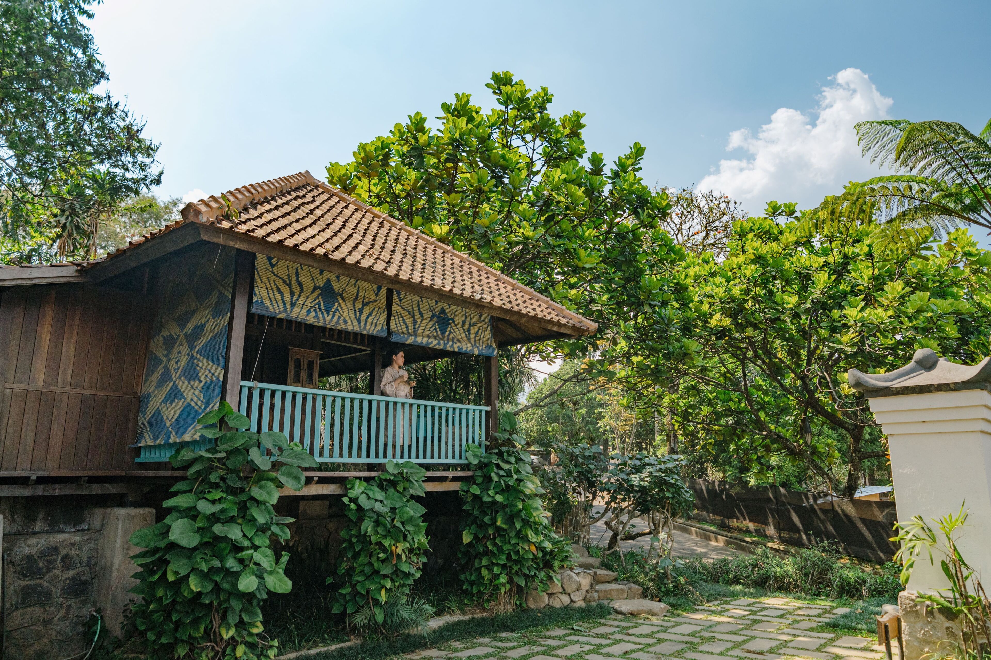 A traditional wooden house with a tiled roof, a balcony, and lush greenery surrounding it.