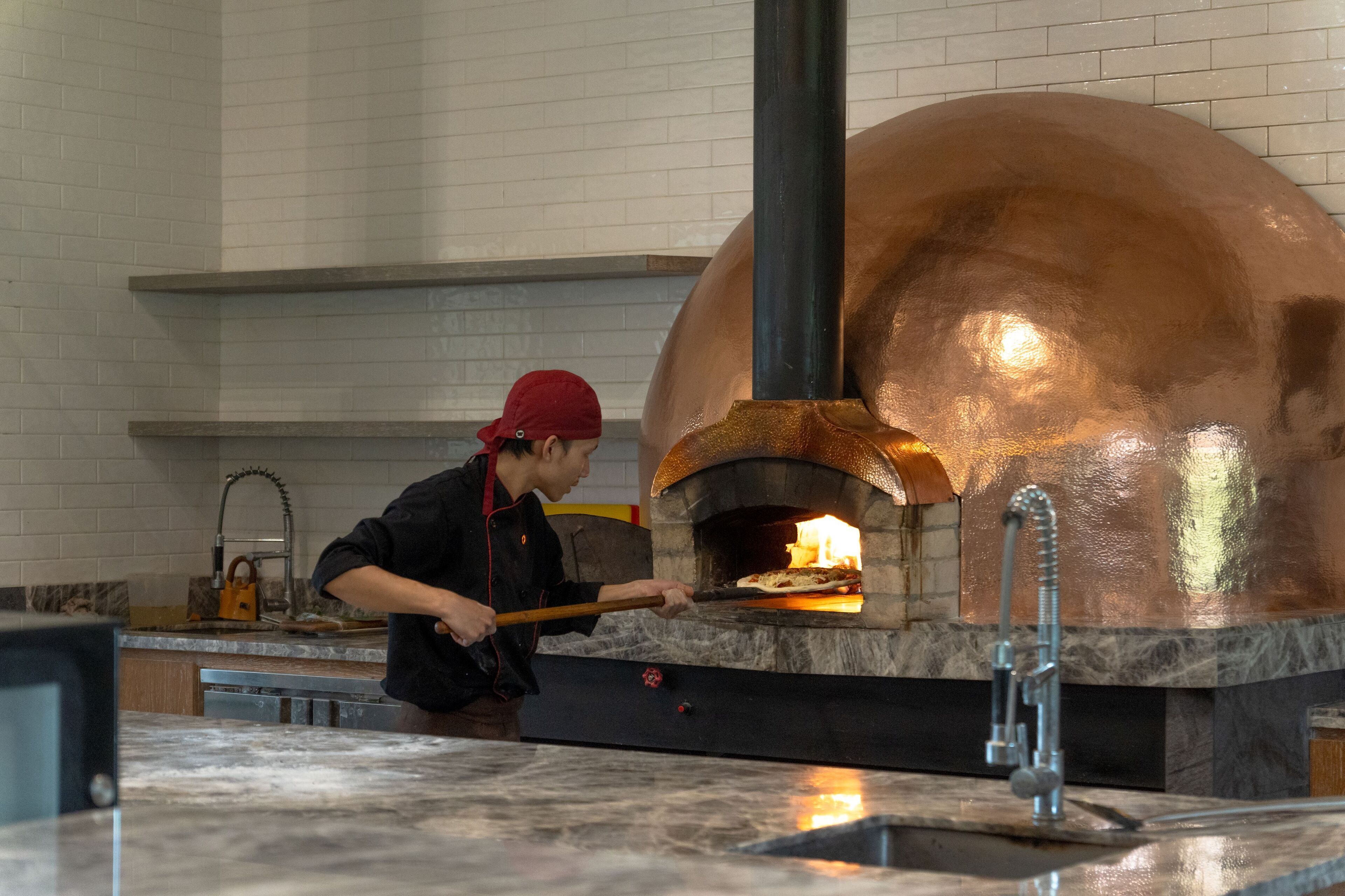 A person using a long-handled tool to manage a pizza inside a large, copper oven.