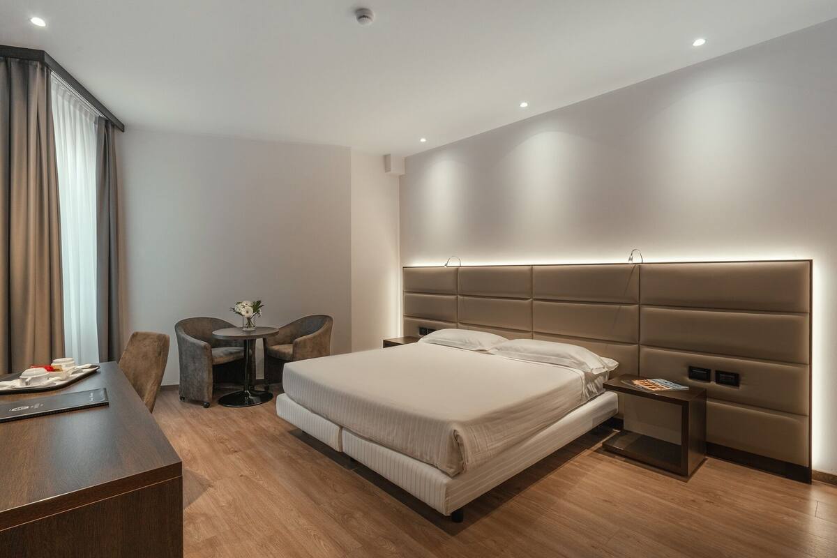 A modern hotel room with a large bed, a desk, a chair, and a small table.