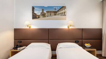 Una camera d'albergo con due letti singoli, un comodino con un telefono e una ciotola di frutta, e un quadro appeso al muro raffigurante un panorama urbano.