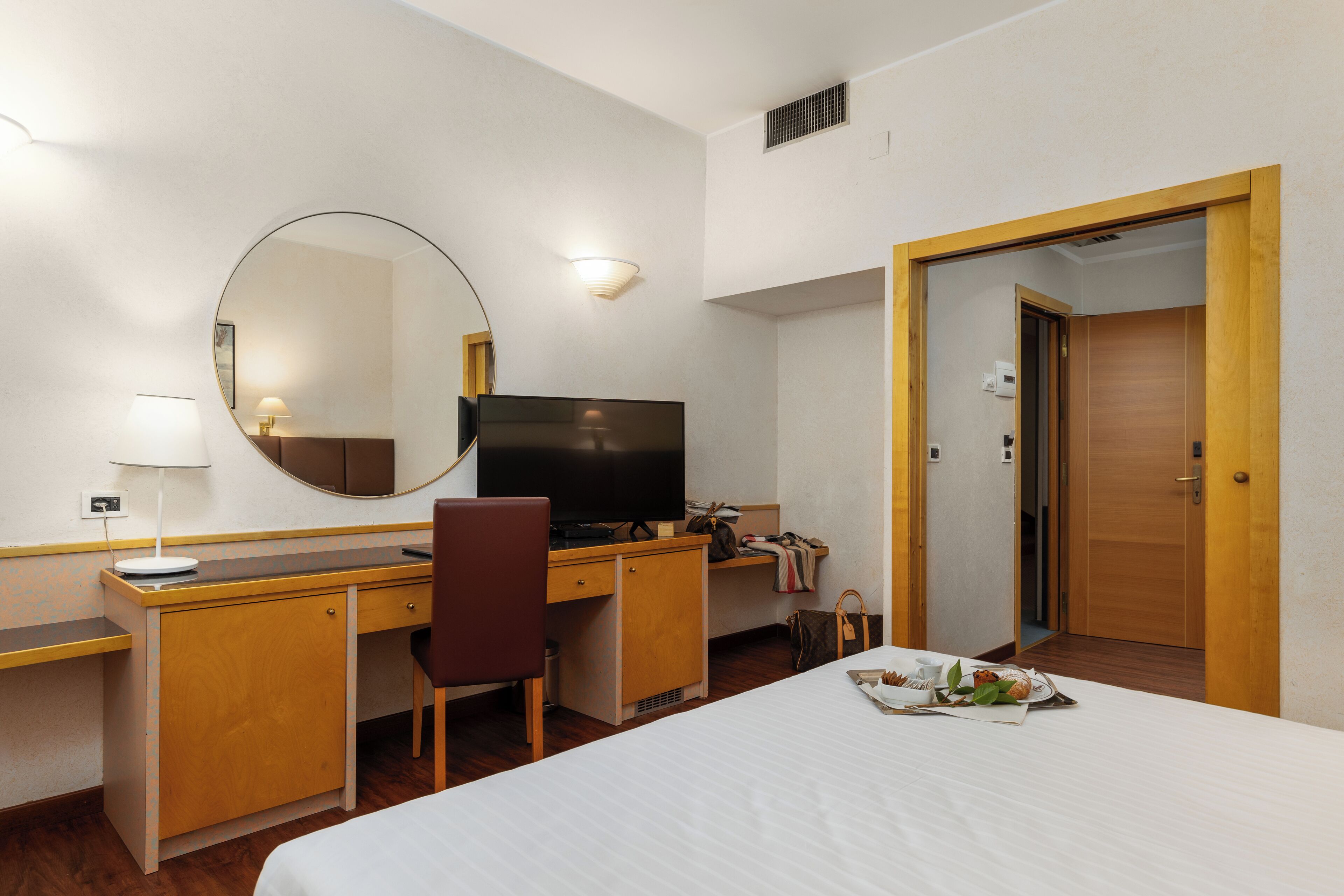Habitación de hotel con cama, escritorio con televisor, silla, espejo y puerta que da a otra habitación.