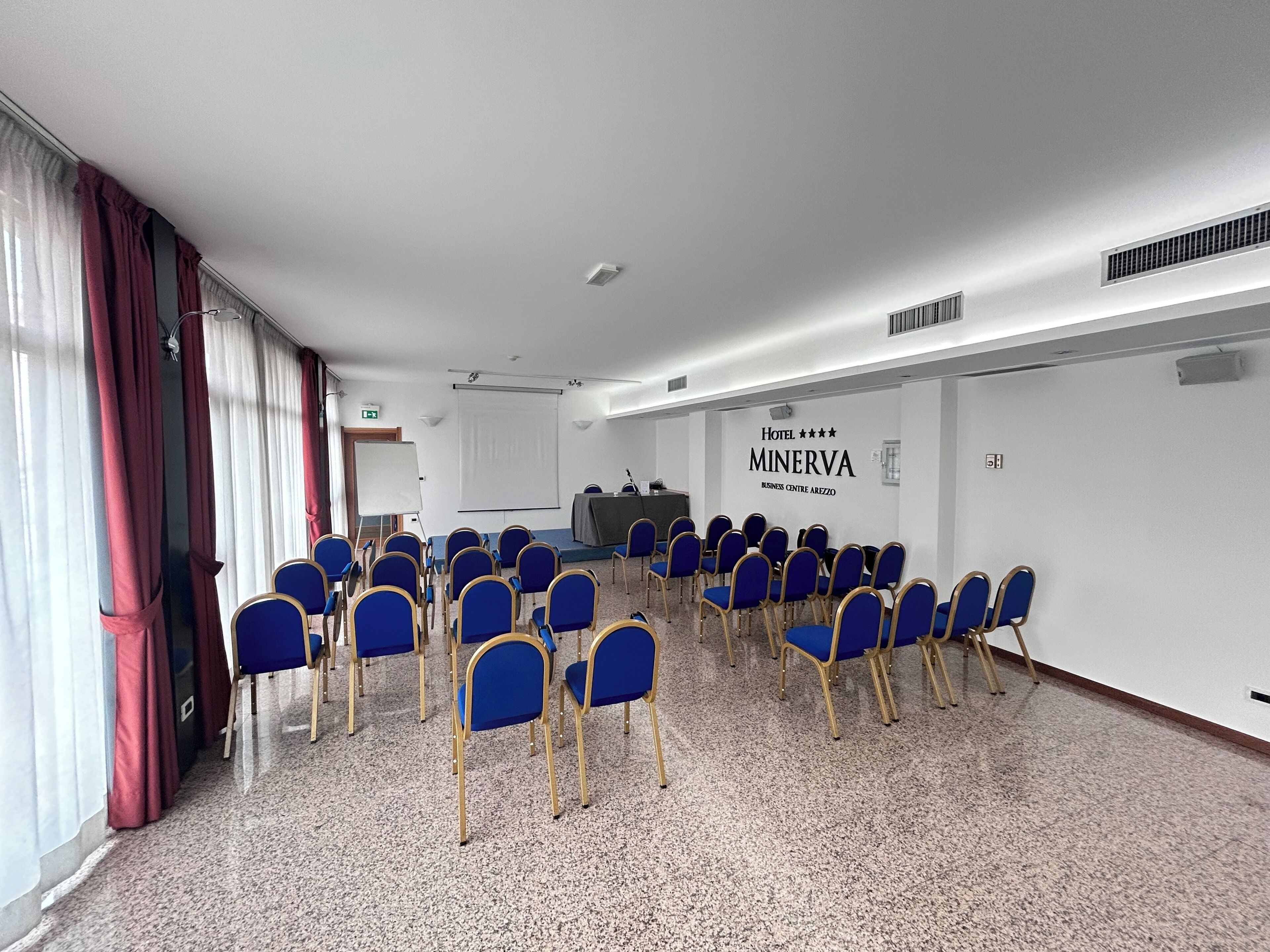 Un salón de conferencias con filas de sillas azules, una pizarra y un cartel del Hotel Minerva.