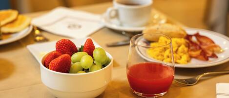 A breakfast table with a bowl of fruit, a glass of red juice, scrambled eggs, bacon, toast, and a cup of coffee.