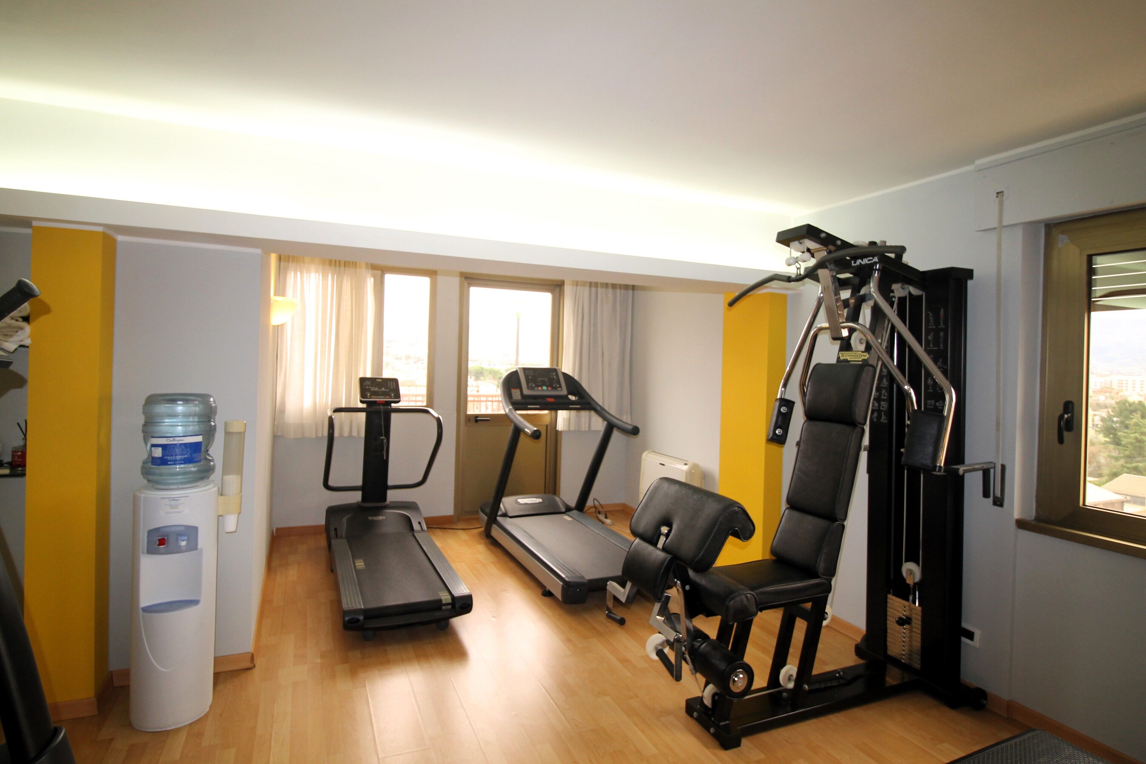Un gimnasio pequeño con una cinta de correr, elíptica y máquina de pesas. Se observa un dispensador de agua. La sala tiene suelo de madera y ventanas grandes con cortinas.