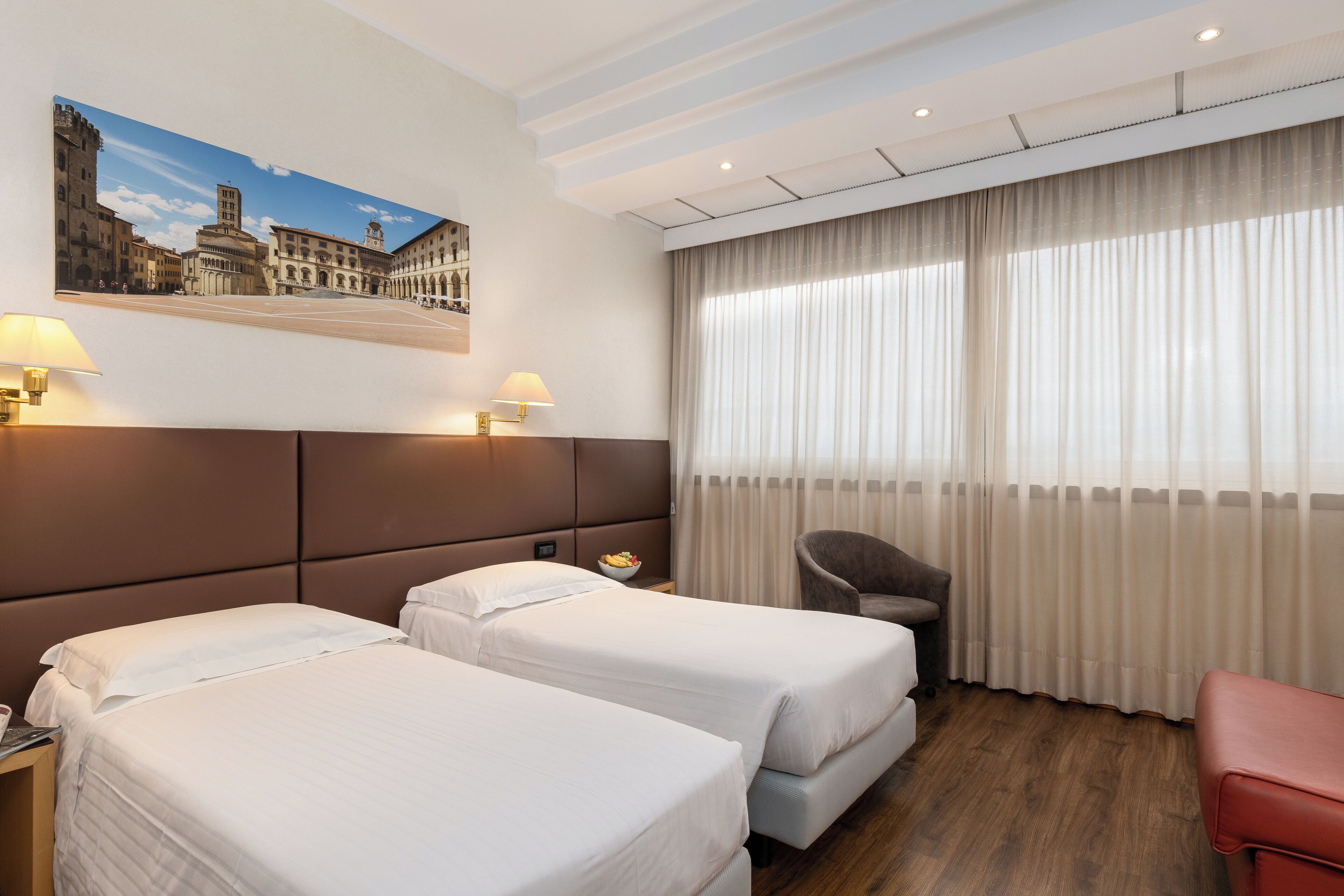 Une chambre d’hôtel avec deux lits, une chaise, un bureau avec une lampe et une grande fenêtre avec des rideaux.
