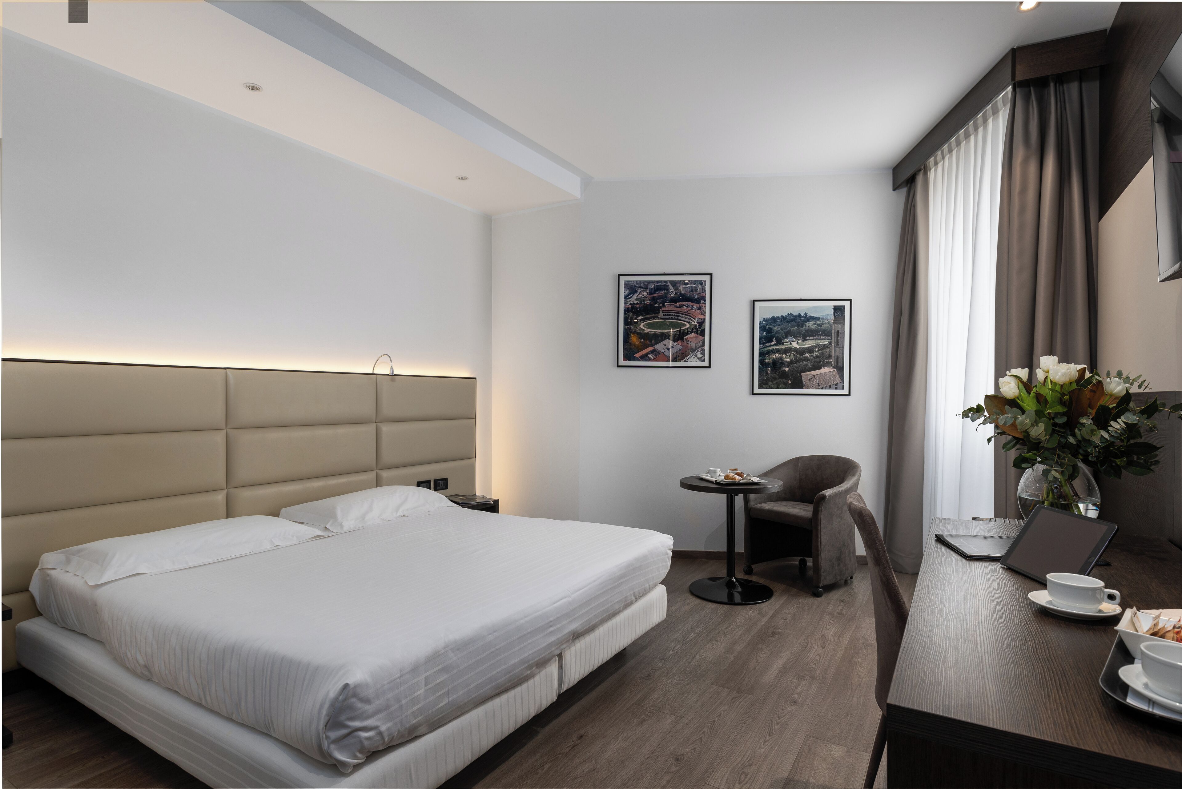 A modern hotel room with a large bed, a small desk with a laptop, a chair, and a vase with flowers.