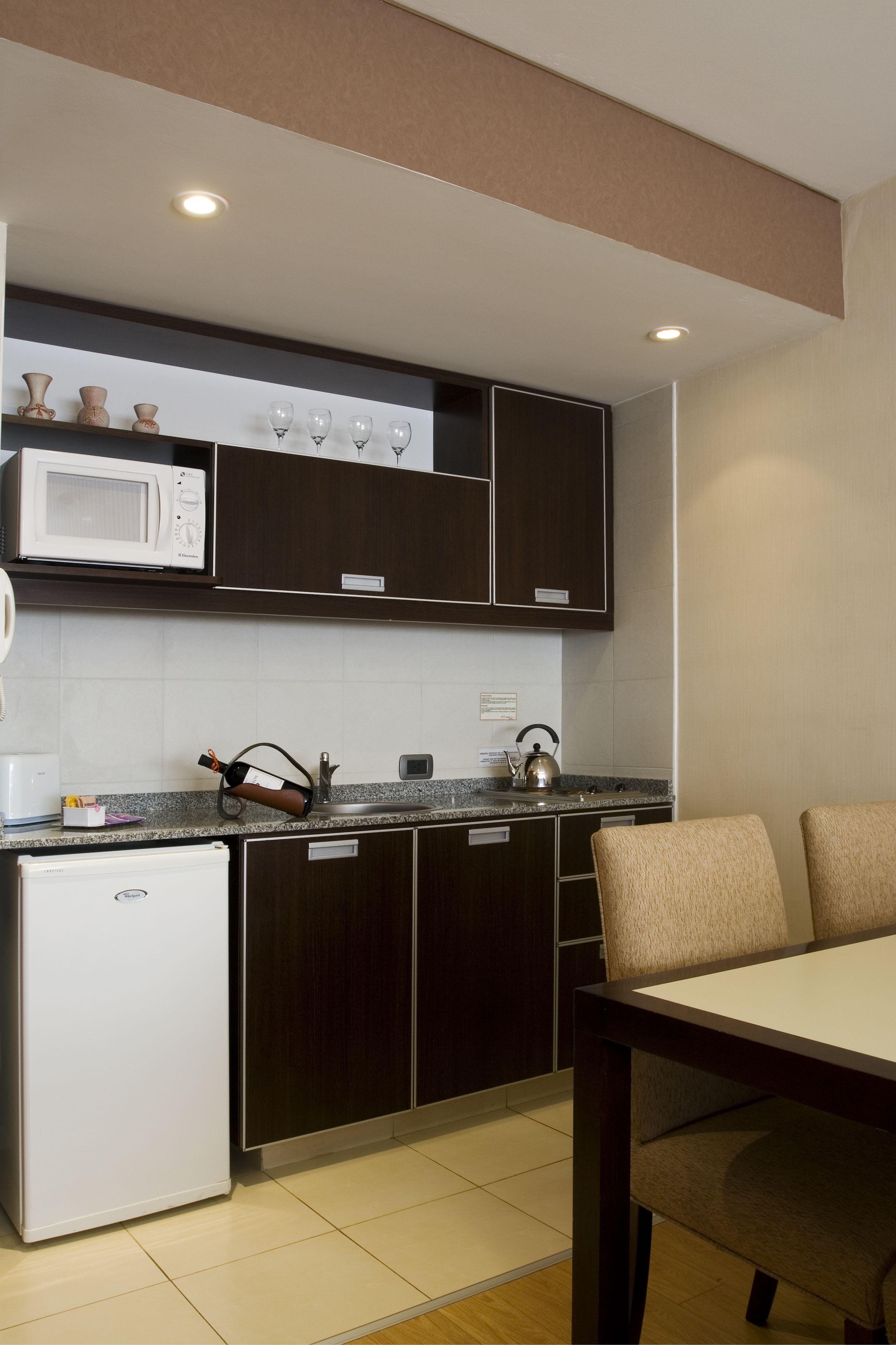 A compact kitchen with dark cabinetry, a microwave, and a kettle on the countertop. There is a small refrigerator and a dining table with chairs.