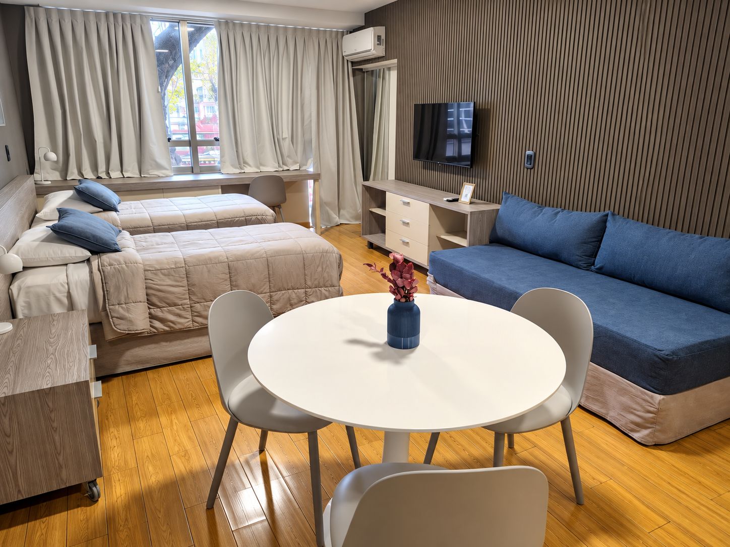 A modern hotel room with a bed, sofa, dining table, chairs, and a TV.