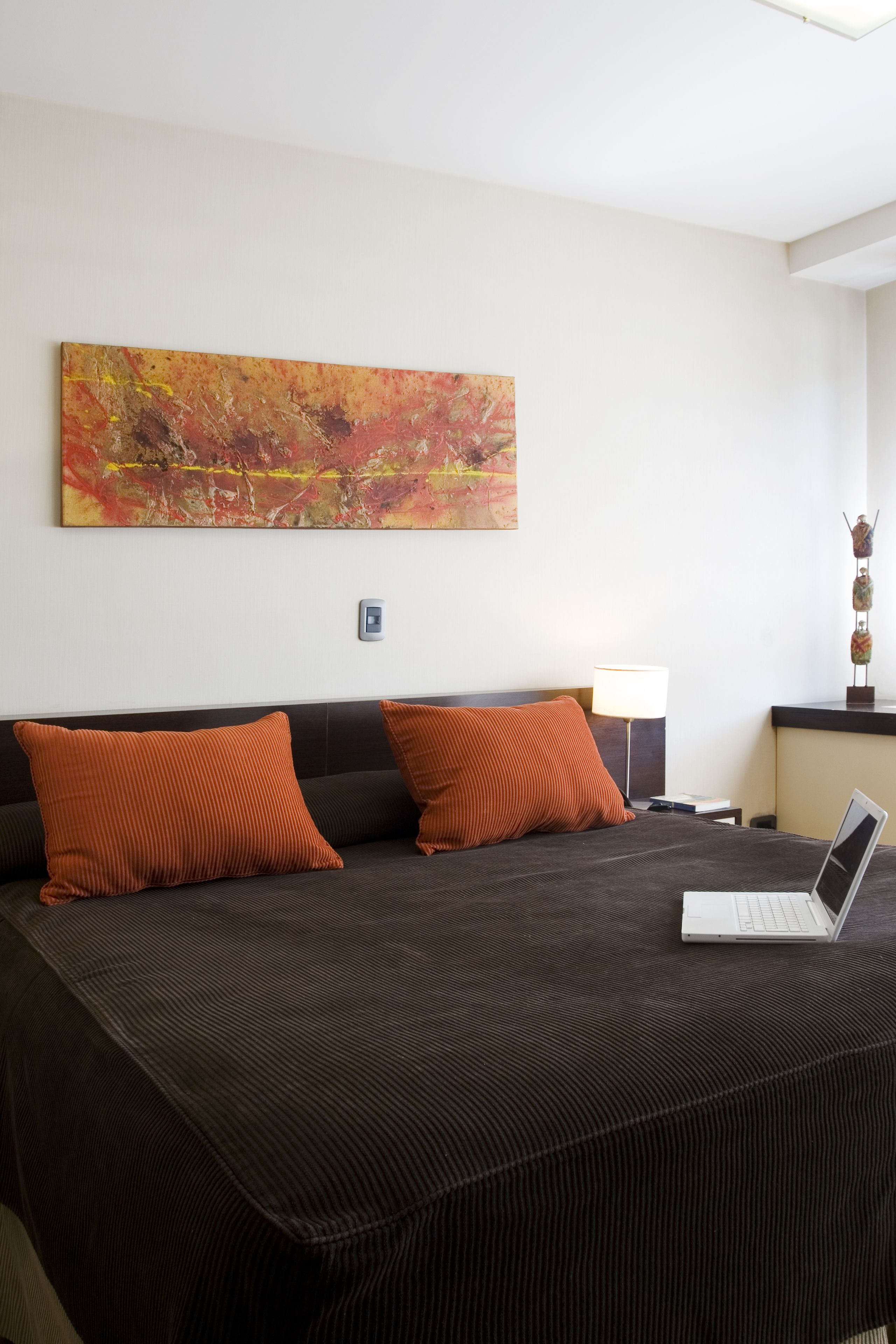 Een hotelkamer met een groot bed, twee oranje kussens, een laptop op het bed, een lamp op een nachtkastje en een kleurrijk abstract schilderij aan de muur.