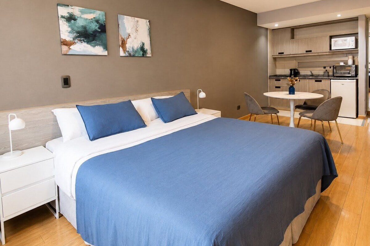 A modern bedroom with a large bed, a bedside table, a desk, and a kitchenette.