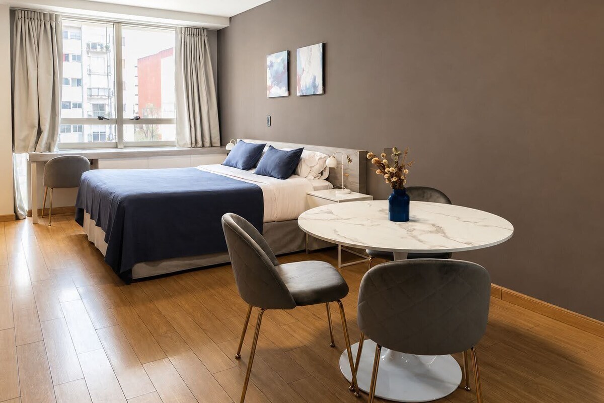 A modern hotel room with a bed, a round table, two chairs, a sofa, and a window with curtains.