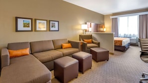 A hotel room with a sofa, ottomans, a chair, a bed, and a desk. There are framed pictures on the wall and a window with curtains.