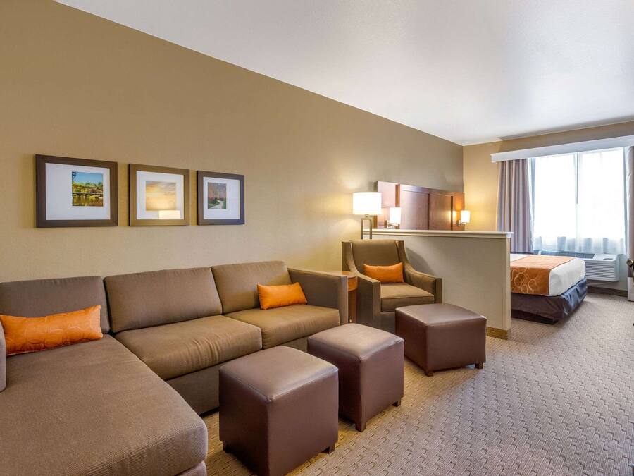 A hotel room with a sofa, ottomans, a chair, a bed, and a desk. There are framed pictures on the wall and a window with curtains.