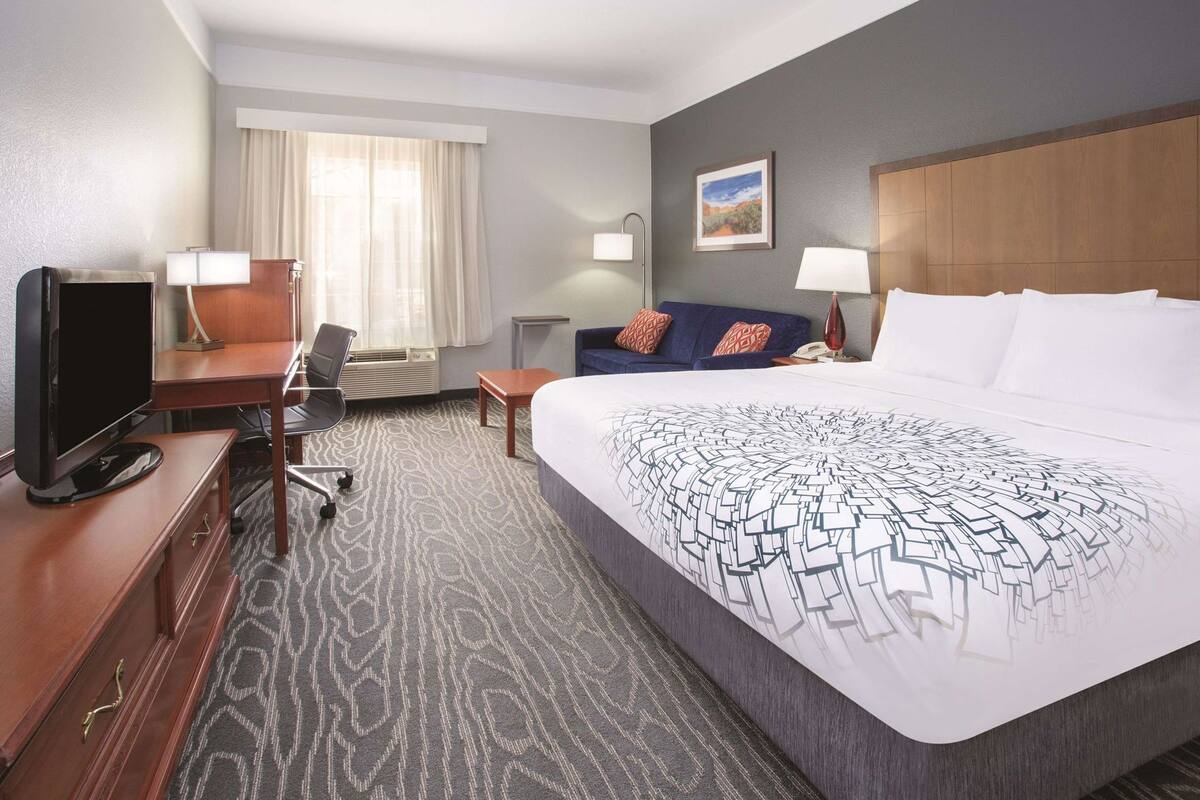A hotel room with a bed, a desk with a TV, a chair, a sofa, and a window with curtains.