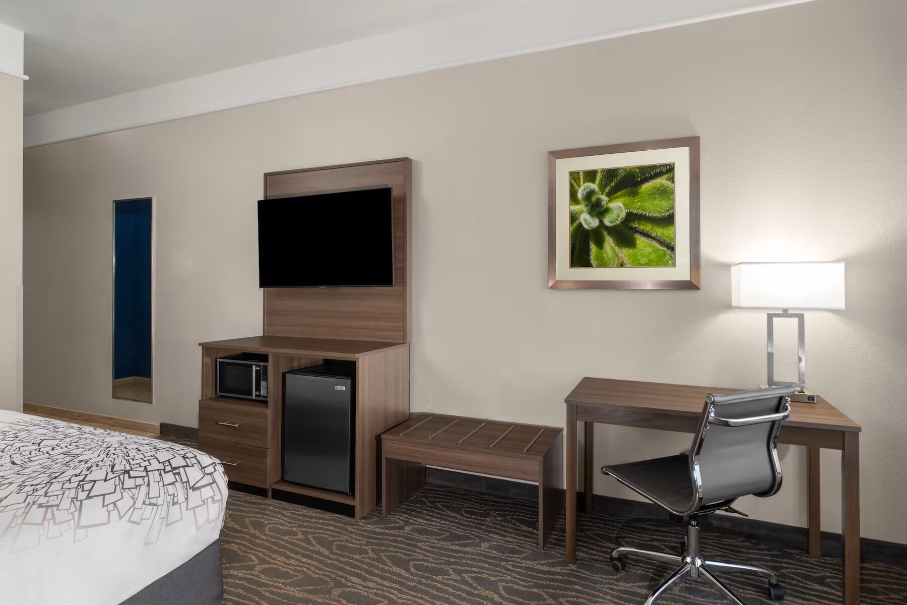 A hotel room with a bed, a desk, a chair, a television, a refrigerator, a lamp, and a framed picture on the wall.