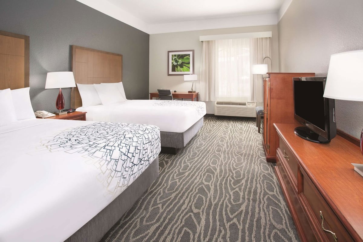 A hotel room with two beds, a television, a desk with a lamp, a window with curtains, and a framed picture on the wall.